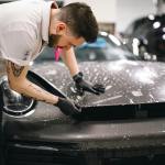 Auto body technician applying protective film on a car hood in a collision repair shop, emphasizing skilled labor and expert techniques in vehicle maintenance.