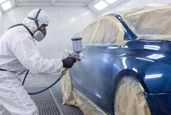 Painter in a protective suit applying a fresh coat of paint to a masked car panel inside a professional spray booth